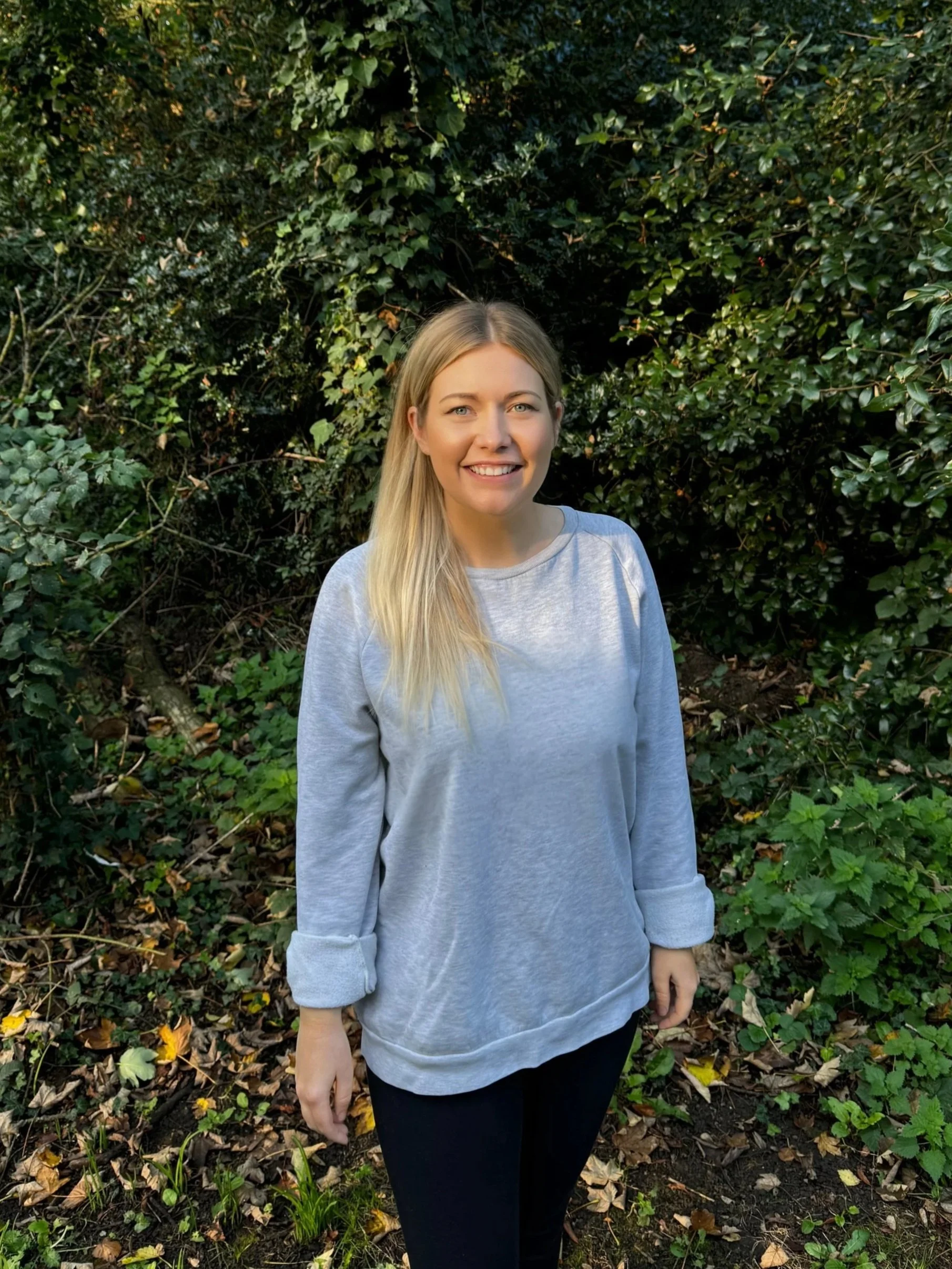 A woman with long blonde hair wearing a light gray sweatshirt standing outdoors in front of dense greenery.