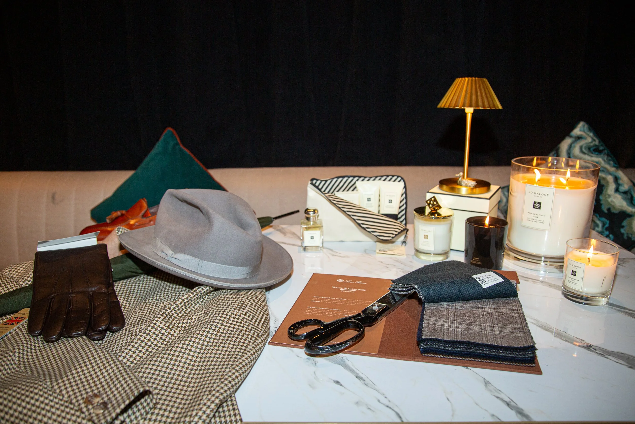 A marble table displaying fabric swatches, scissors, a gray hat, gloves, perfume bottles, candles, and a striped jewelry box, with a sofa and pillows in the background.