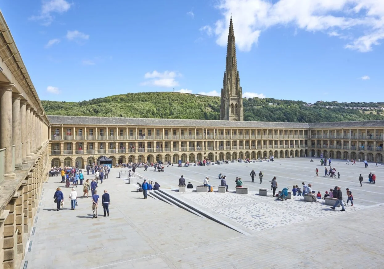 People walking and sitting in an open courtyard of a historic building with a large spire church in the background, surrounded by green hills.