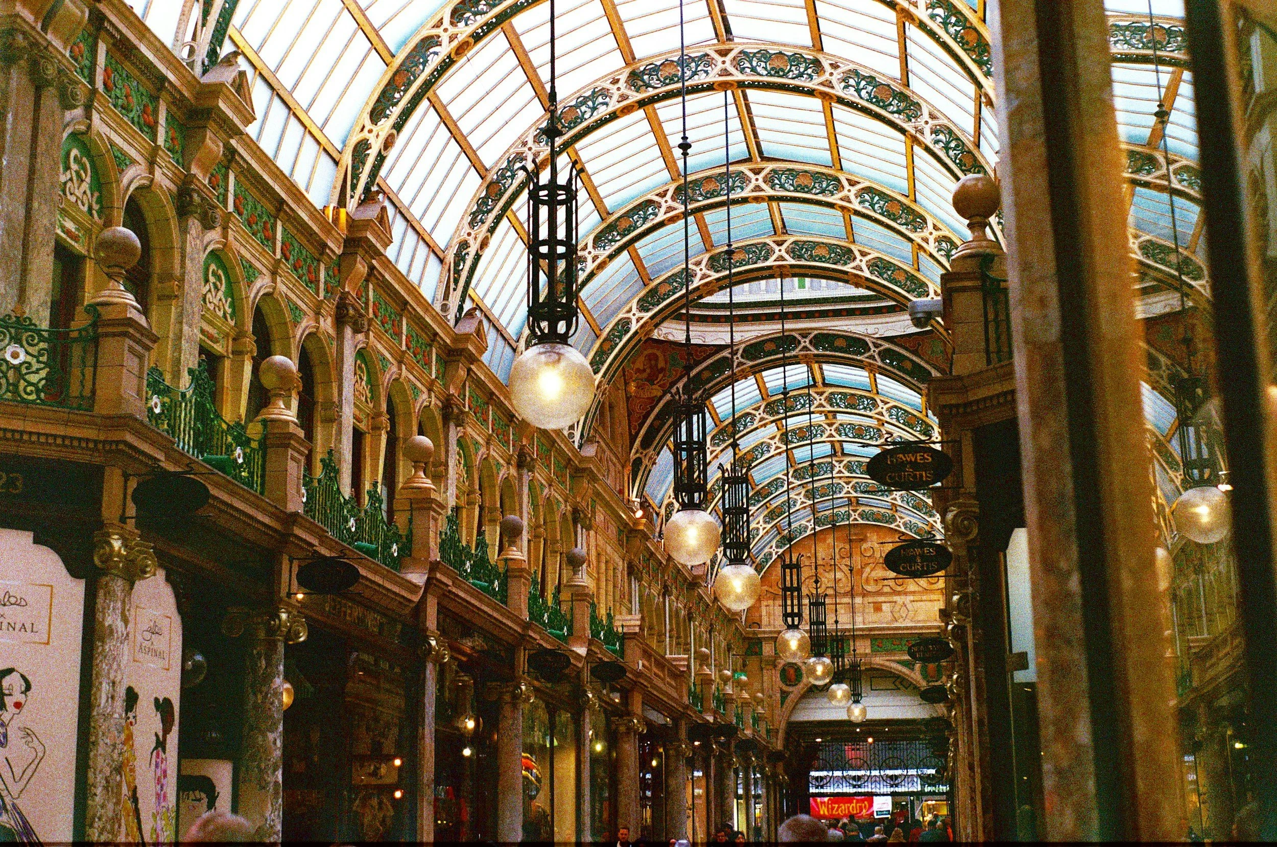 Interior of a shopping arcade with ornate arching glass ceilings, vintage-style pendant lighting, decorative ironwork, and storefronts on either side.