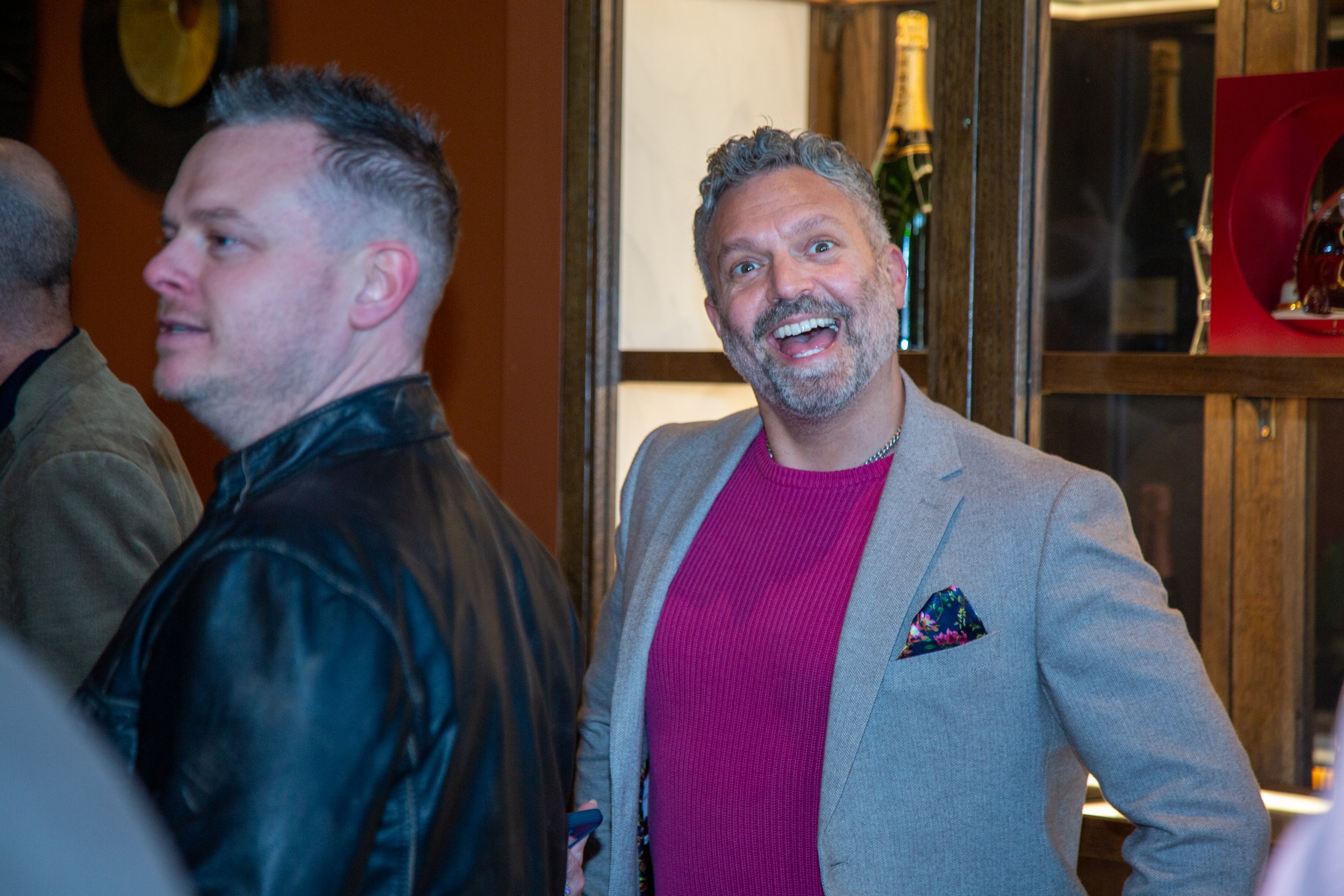 A man with gray hair and a beard, wearing a light gray blazer, pink sweater, and a colorful pocket square, smiling enthusiastically in a social setting. Another man with short hair and a black leather jacket is in the foreground, partially turned awa