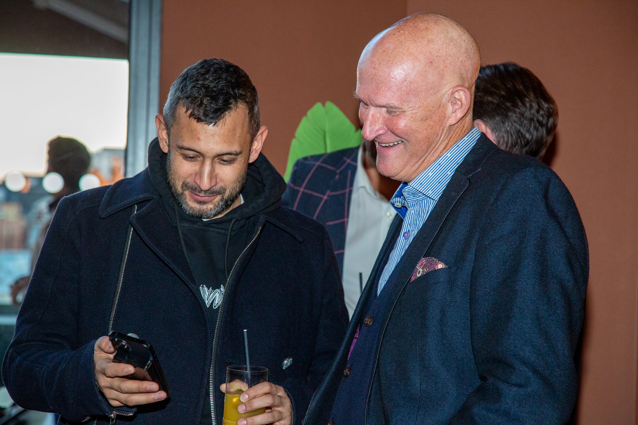 Two men are smiling and looking at a smartphone at a social event. One is holding a glass of orange drink with a straw.