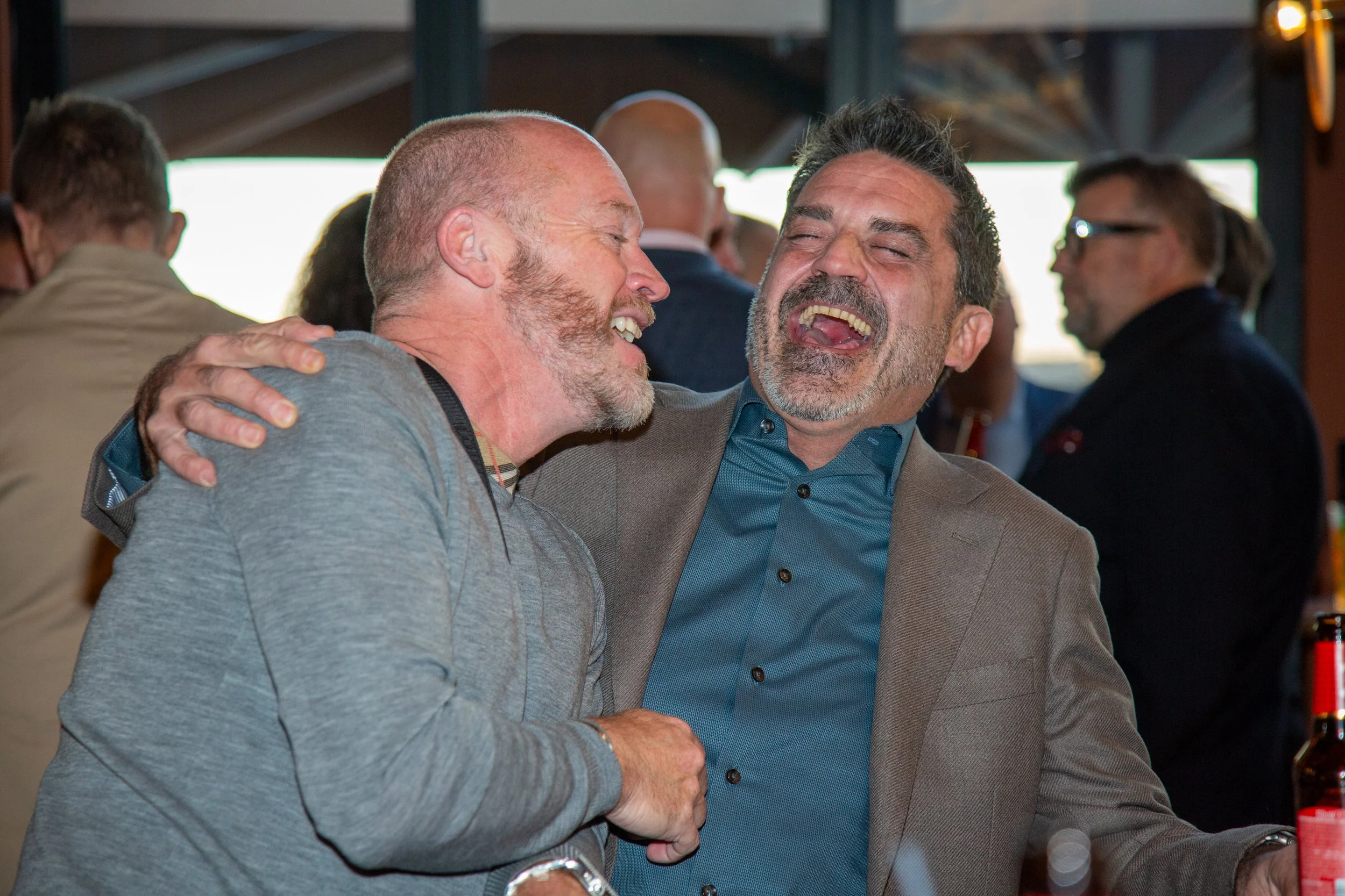 Two middle-aged men laughing and hugging at a social gathering, with other people in the background in a casual setting.