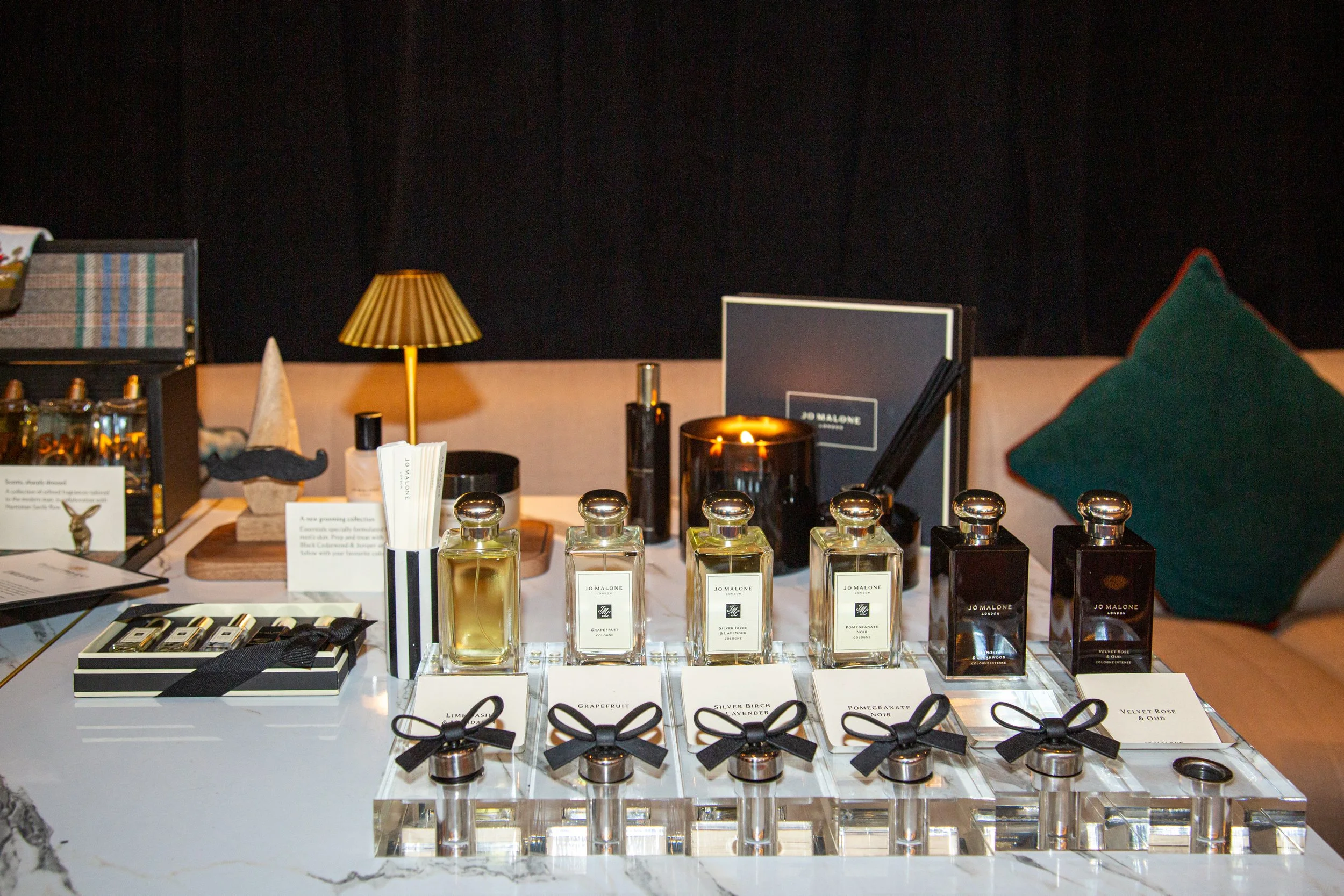 Display of Jo Malone perfumes and scented products arranged on a white marble table, with a black background and a golden lamp behind.