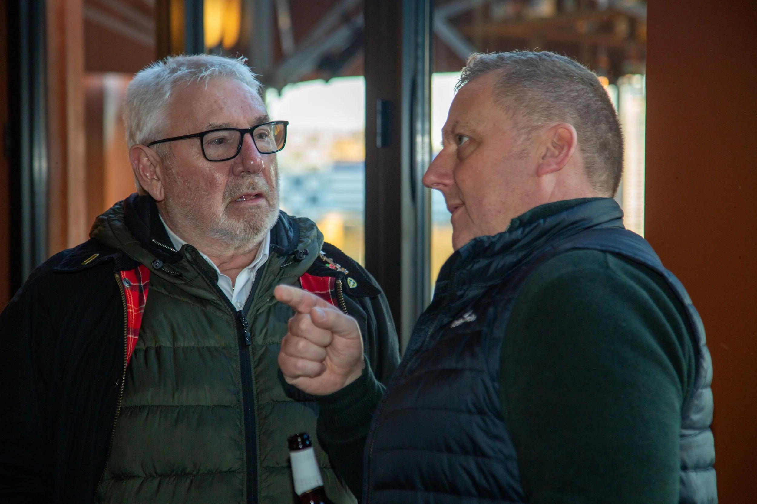 Two men engaged in a serious conversation indoors, one with gray hair and glasses, the other with short dark hair, both wearing jackets.
