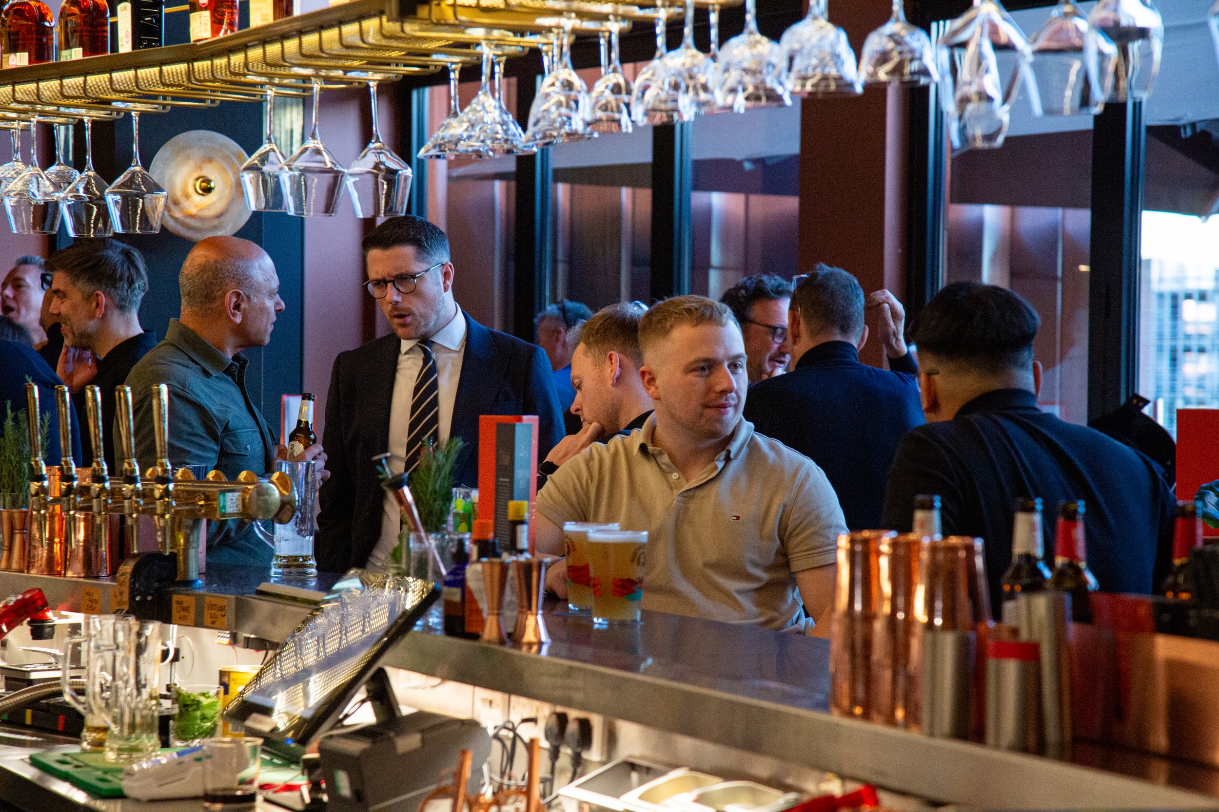 People socializing at a bar with drinks and glasses hanging overhead