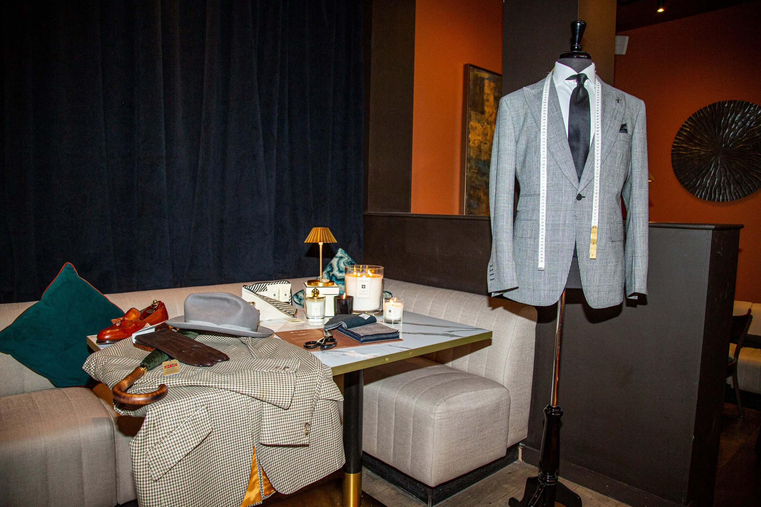 A collection of men's clothing and accessories on a table and mannequin, including a tweed blazer, hat, umbrella, and shoes, in a restaurant setting.
