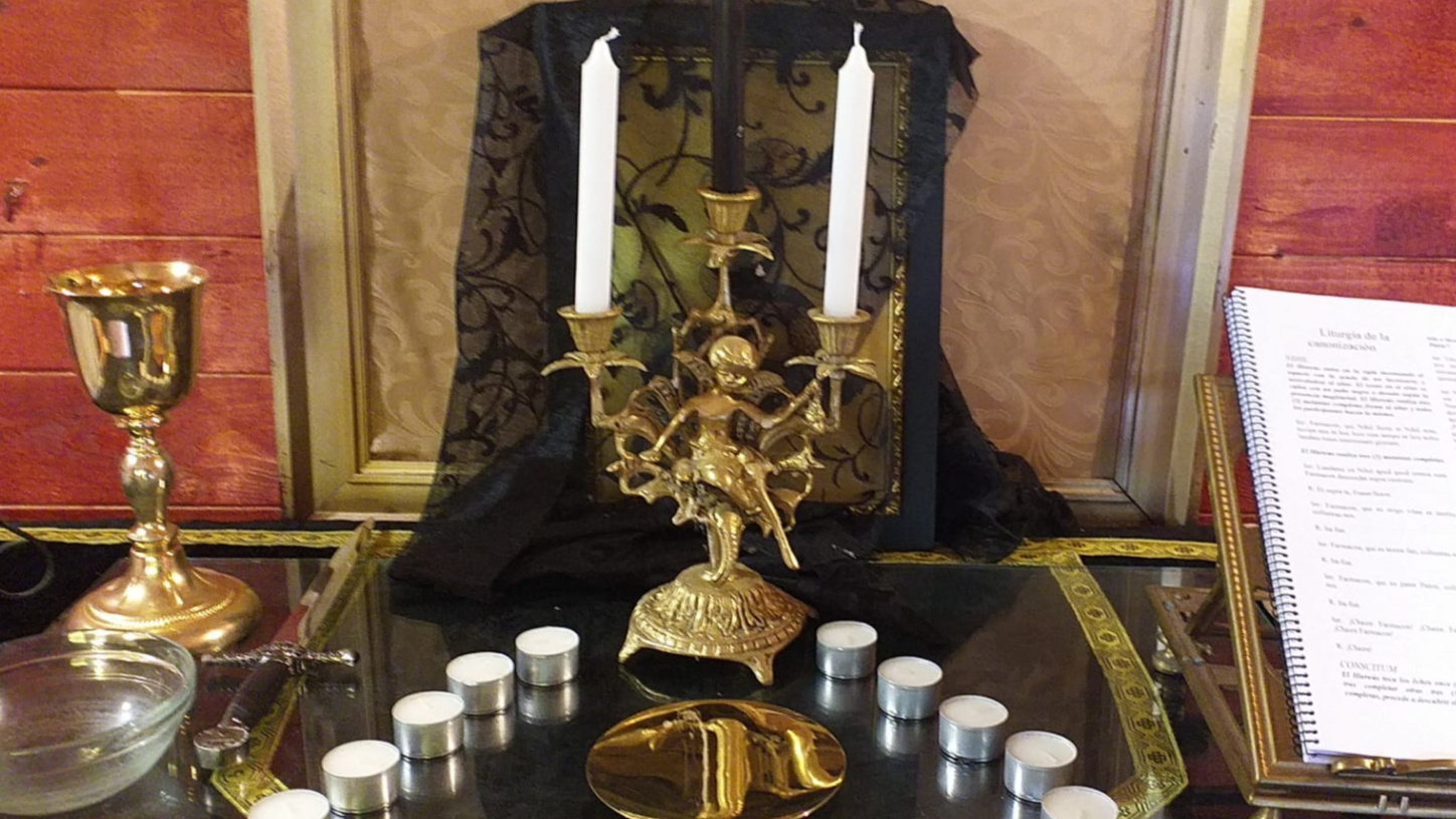 Gold vintage candelabrum with three white candles, surrounded by small tea light candles, set on a table with a decorative black and gold border, with a prayer book and a brass goblet in the background.