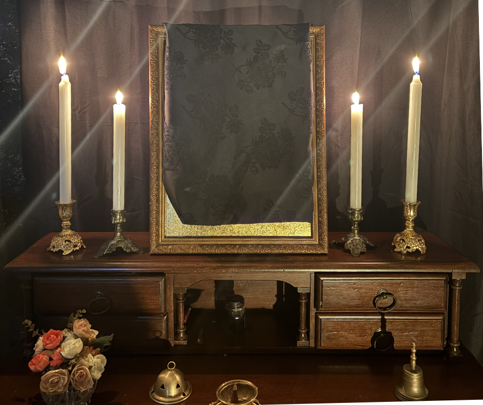 A wooden table holding four lit candles with ornate ancient brass candle holders, a Serpent golden icon covered with a black cloth, a bouquet of roses, and various small decorative items in a dimly lit room.