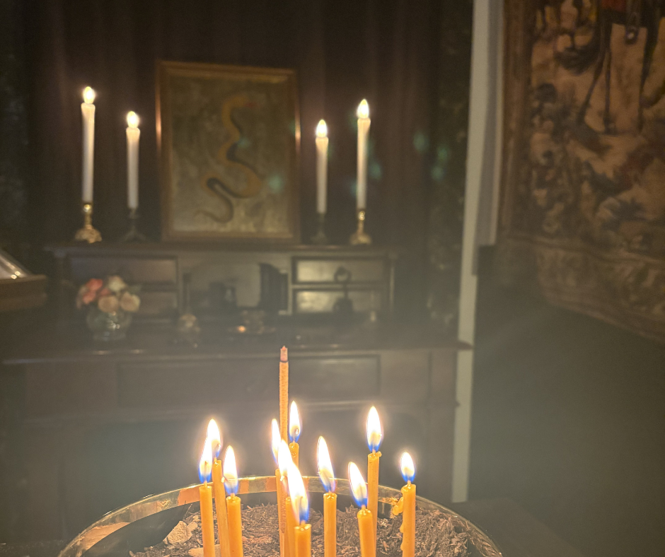A black and gold bowl containing eleven small devotional candles in the foreground and a dark room with a liturgical altar, containing four more big candles and a Serpent icon in the background.
