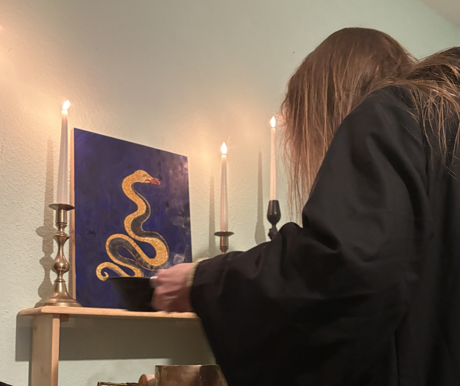 Person with long hair  burning incense in front of a Serpent Icon on the wall, with three lit candles on candle holders nearby.