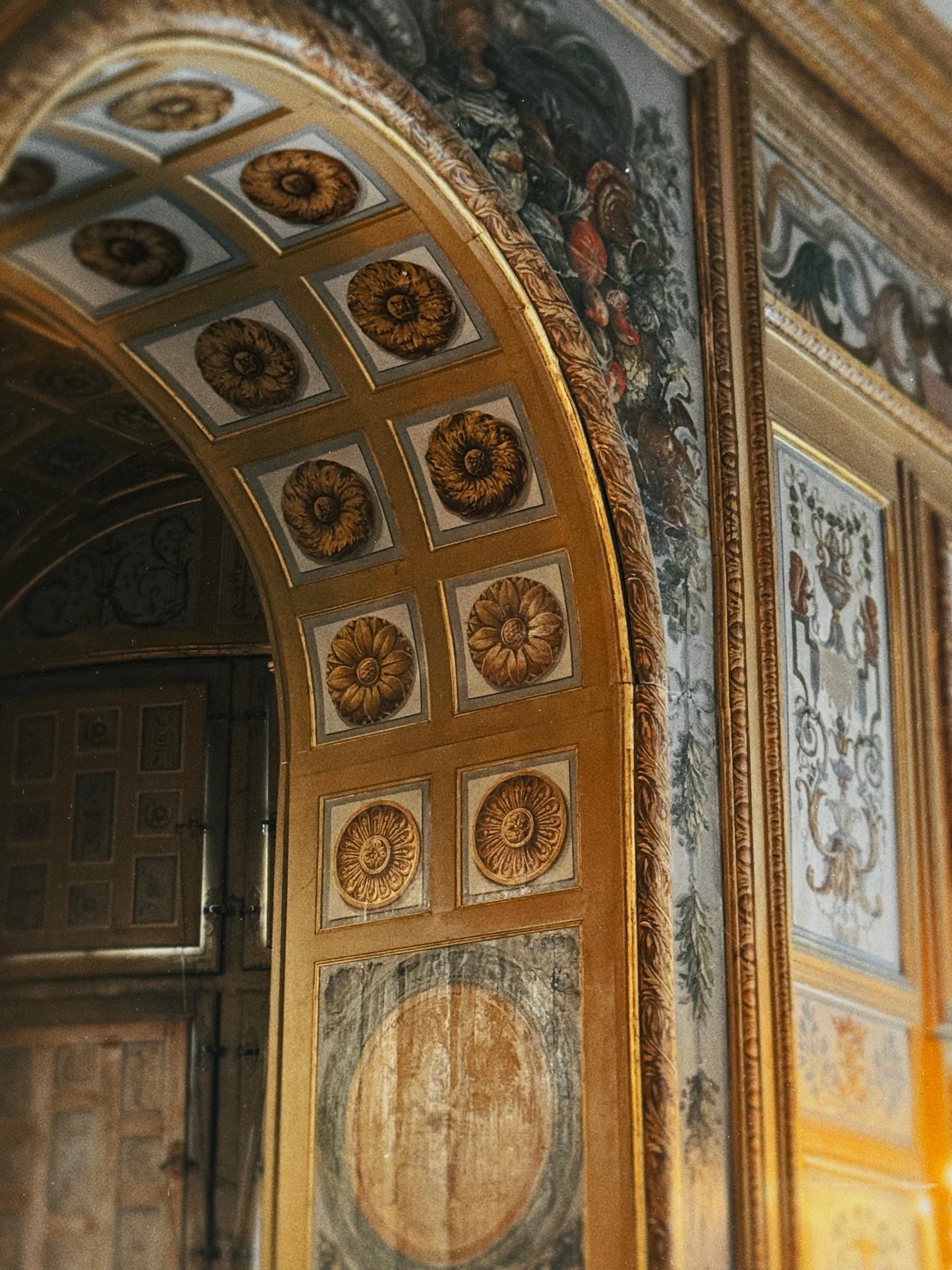 An ornate, decorative ceiling with gold accents, floral motifs, and intricate molding, featuring a painted scene with figures and animals.