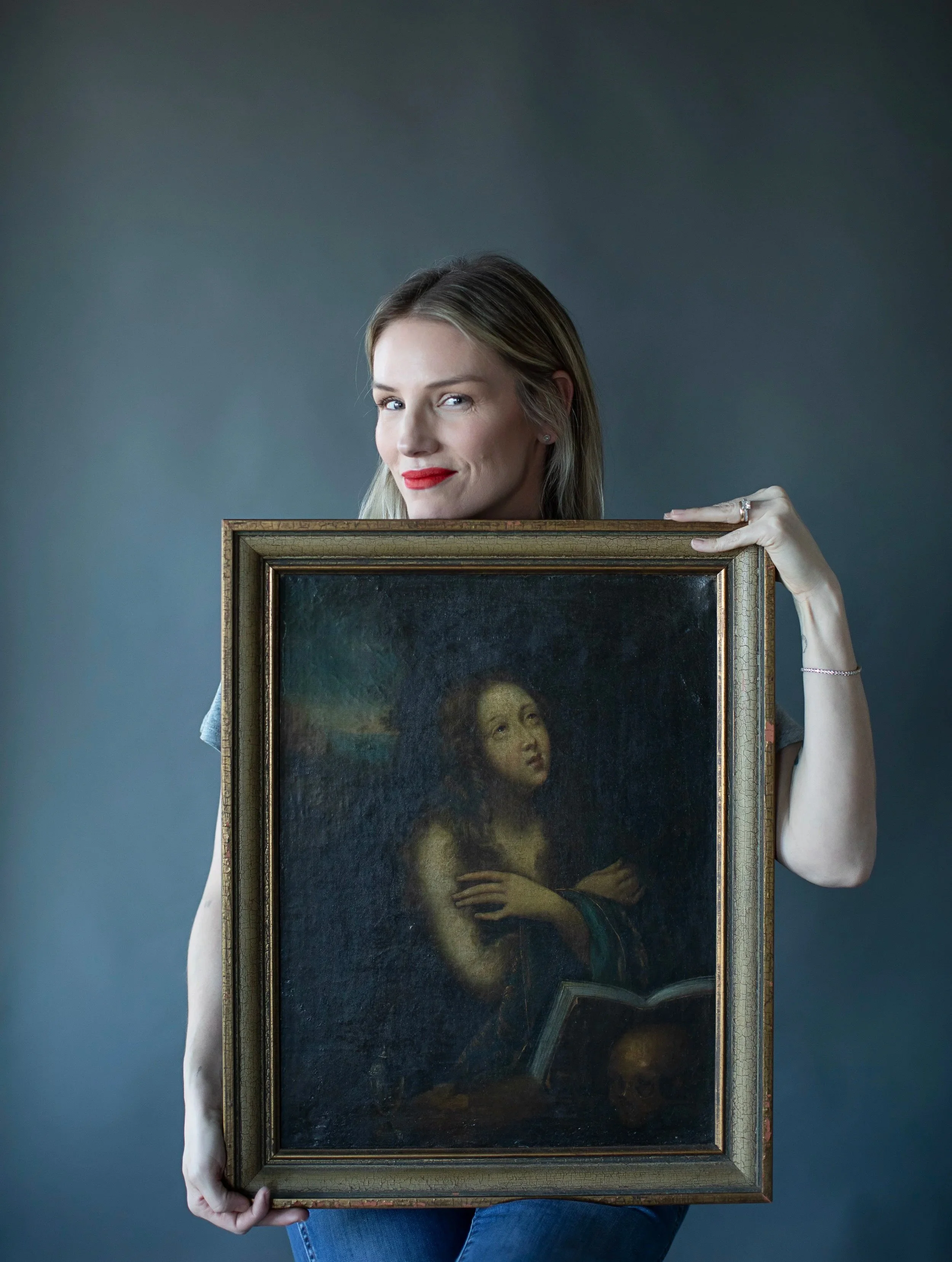 A woman with blonde hair and red lipstick holding a framed painting of the Mona Lisa in front of her body, obscuring her torso.