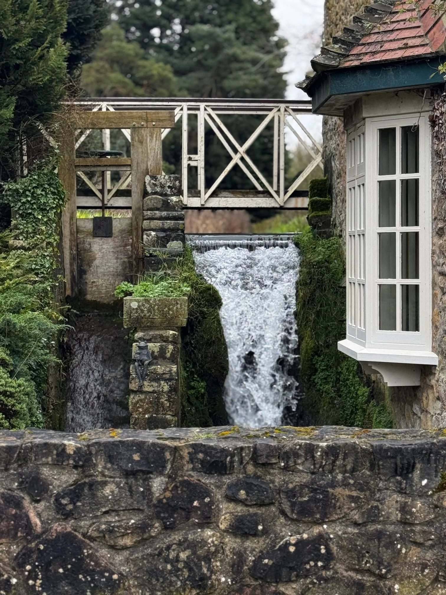 A small waterfall or stream flowing through a stone channel, with a white wooden bridge and a white windowed house in the background, all surrounded by greenery.