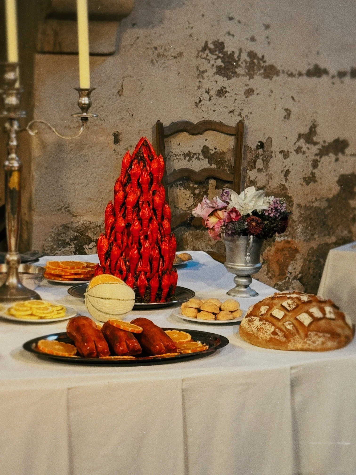 A table set with various foods including a cake shaped like a bunch of red peppers, sausages with orange slices, a loaf of bread, and several plates of other snacks, with a decorative candelabrum and a floral arrangement in the background.