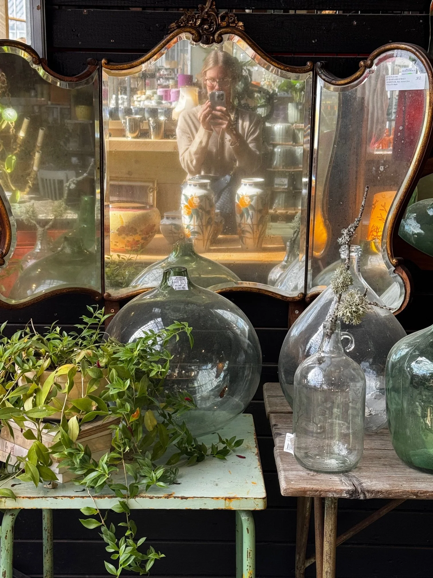 A person taking a photo of a vintage mirror with a reflection of her holding a camera. The mirror is surrounded by various glass vases and ornaments on a display table in a store.