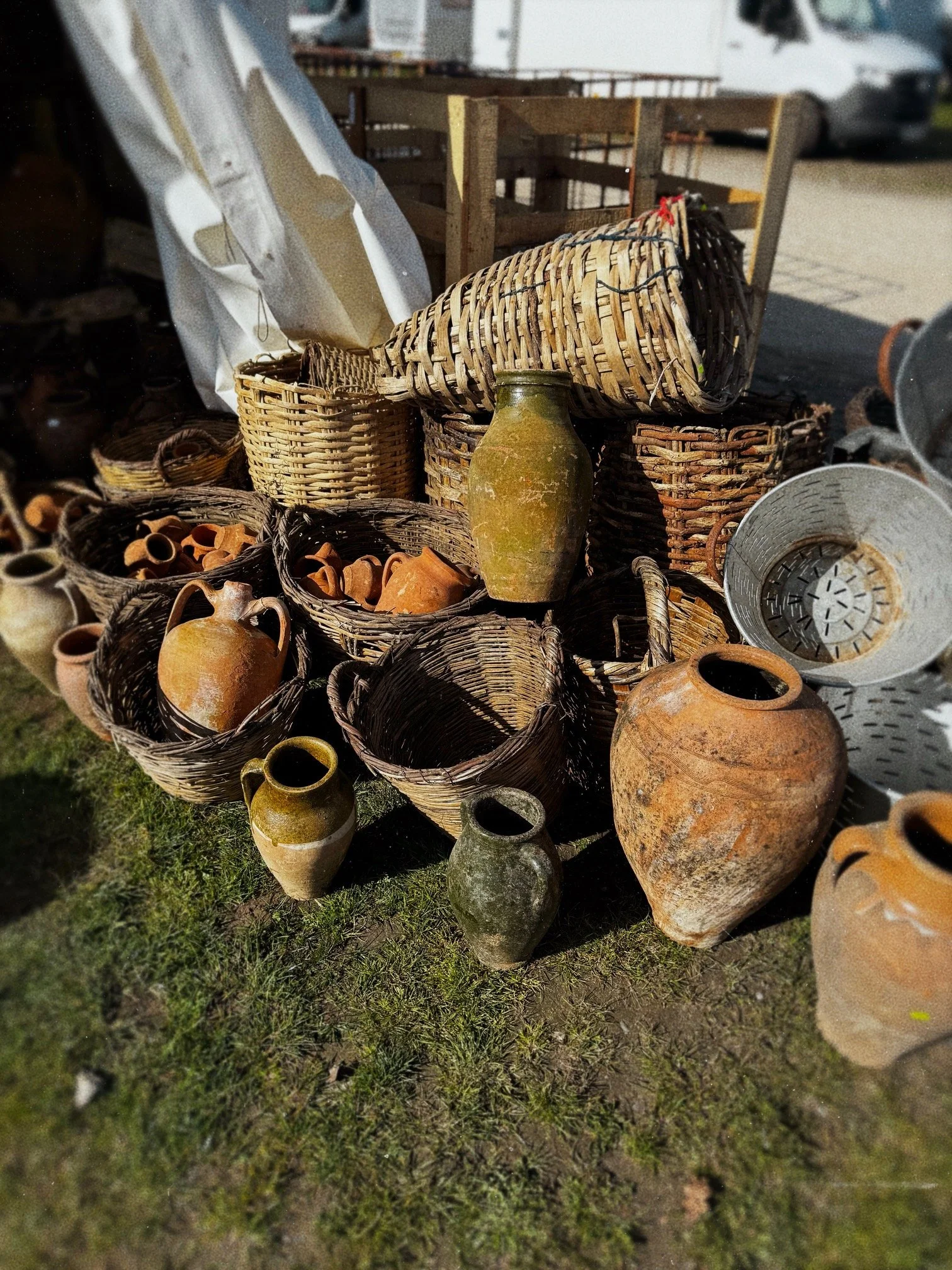 Table filled with various old clay pots, baskets, and woven containers outdoors on grass, with wooden crates and a white vehicle in the background, illuminated by sunlight.