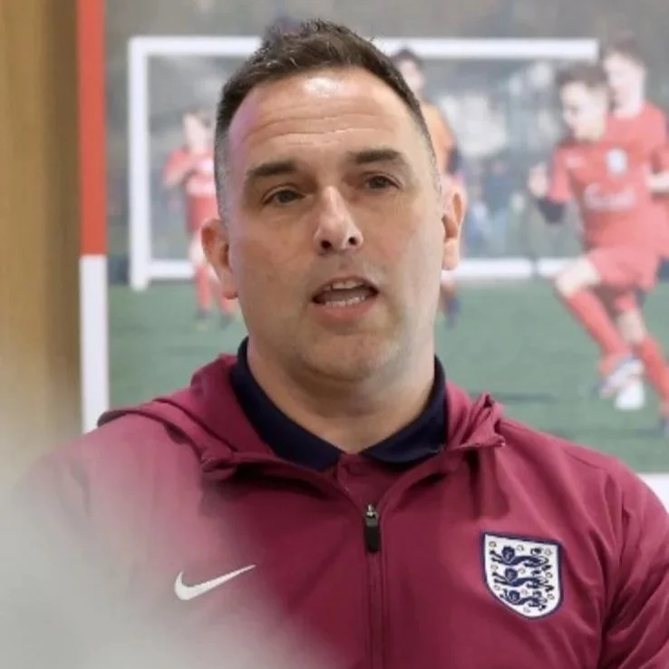 A man in an England football team jacket speaking indoors, with a blurred background of a soccer photo with players in red kit.