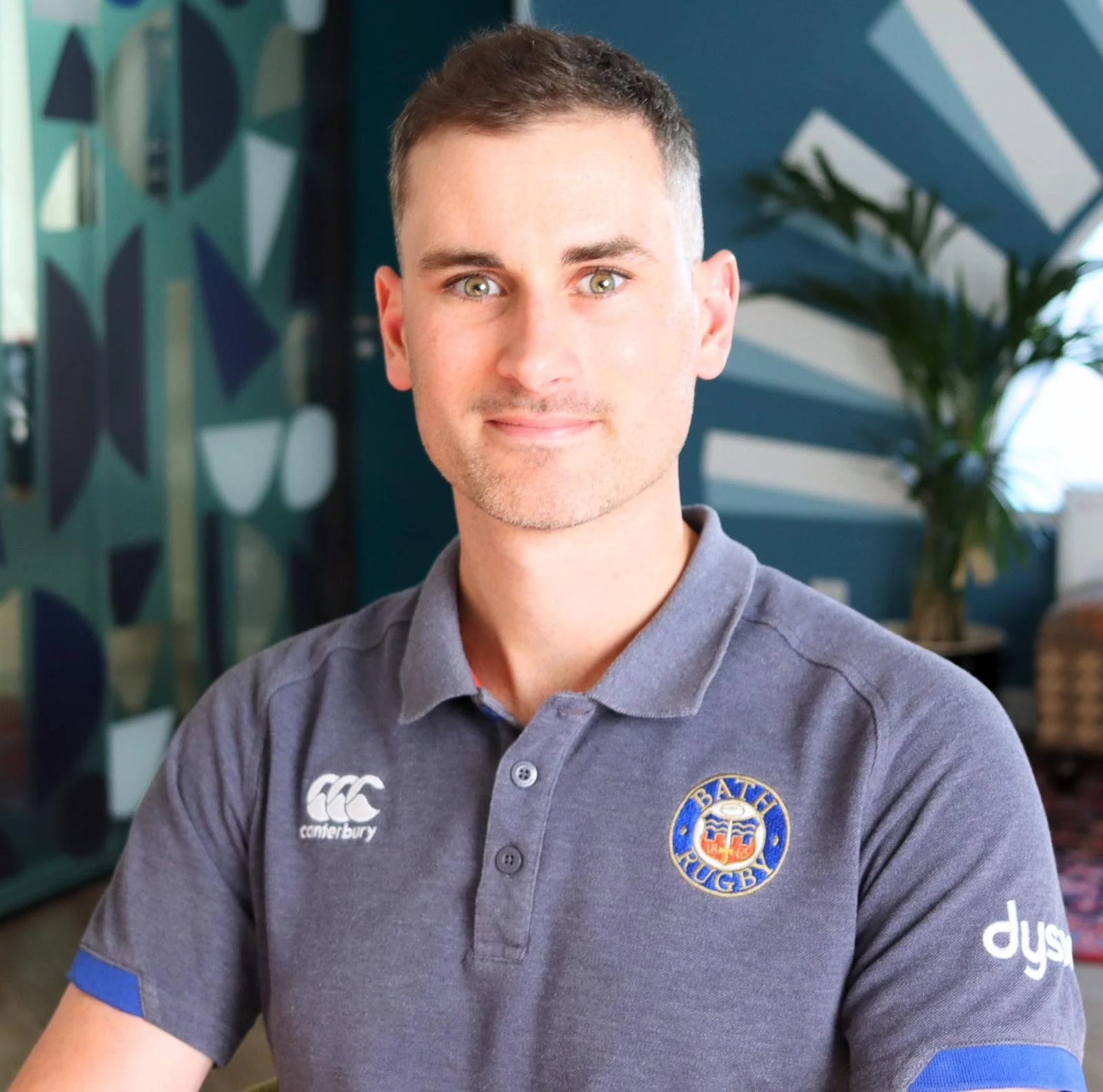 Portrait of a young man with short dark hair and green eyes wearing a grey uniform shirt with logos, sitting in an indoor setting with abstract wall art and a plant in the background.