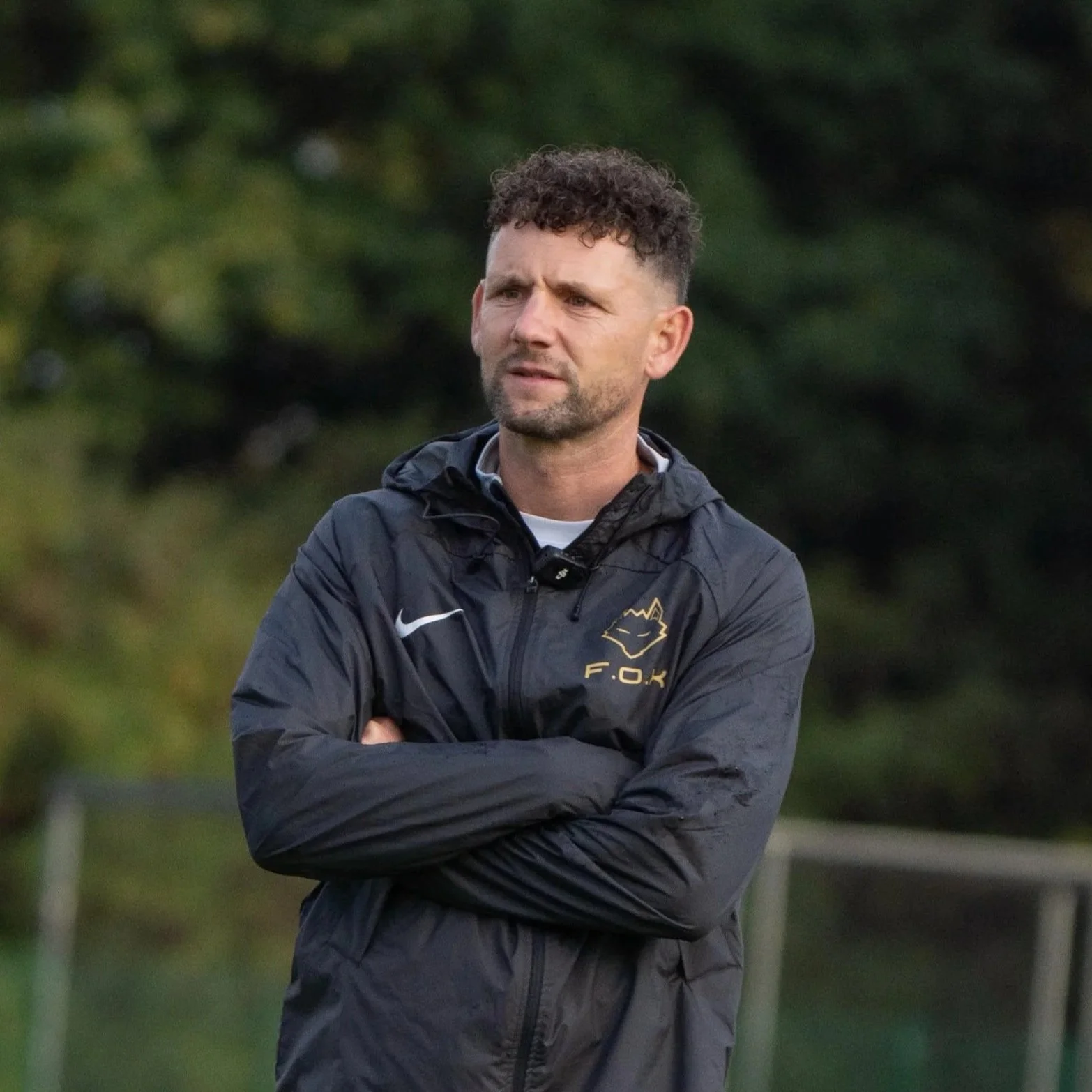 A man with short curly hair and a beard stands outdoors with arms crossed, wearing a dark Athletic jacket with logo of a fox and the initials F.O.K. on it, against a background of green trees.