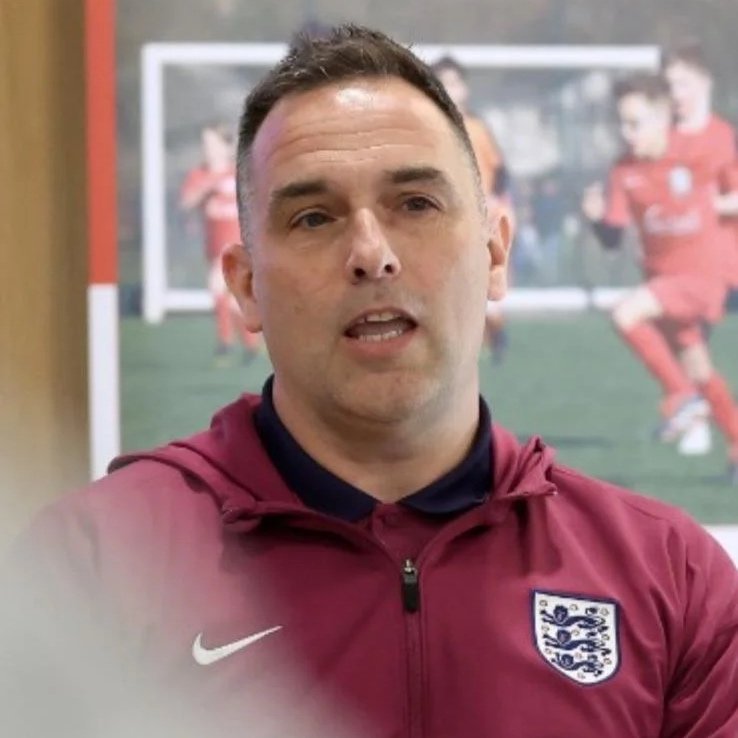 A man wearing a maroon Nike jacket with the England football badge, standing in front of a blurred soccer game poster.