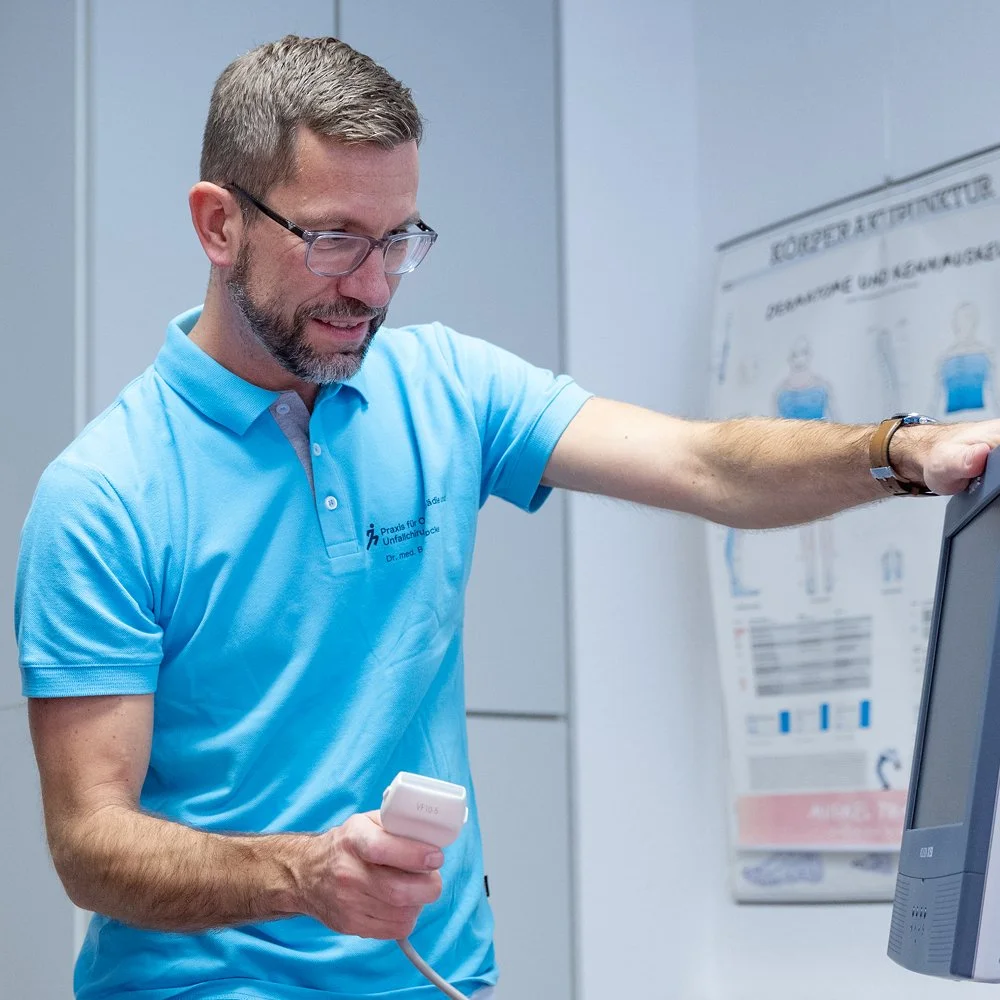 Ein Mann mit Brille und kurzhaarigem Bart in einem blauen Polo-Mann-Shirt, der ein medizinisches Gerät in der Hand hält und einen Bildschirm berührt, vermutlich in einer medizinischen Praxis oder einem Gesundheitszentrum.