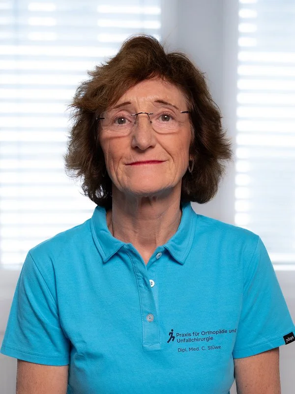 Ältere Frau mit Brille und lockigem braunem Haar, trägt eine blaue Pflegeuniform mit Logo und Text, im Hintergrund Fenster mit geschlossenen weißen Jalousien.