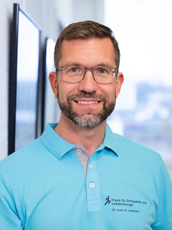 Ein Mann trägt ein hellblaues Polo-Shirt mit einem Logo und Text, der auf eine orthopädische Praxis und Chirurgie hinweist, und steht vor einem Fenster mit Stadtblick.