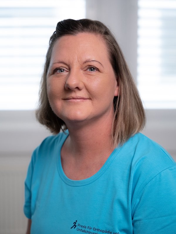 Eine Frau mit braunen Haaren, blauer T-Shirt, vor einem Fenster mit weißen Jalousien, lächelnd in die Kamera schauend.