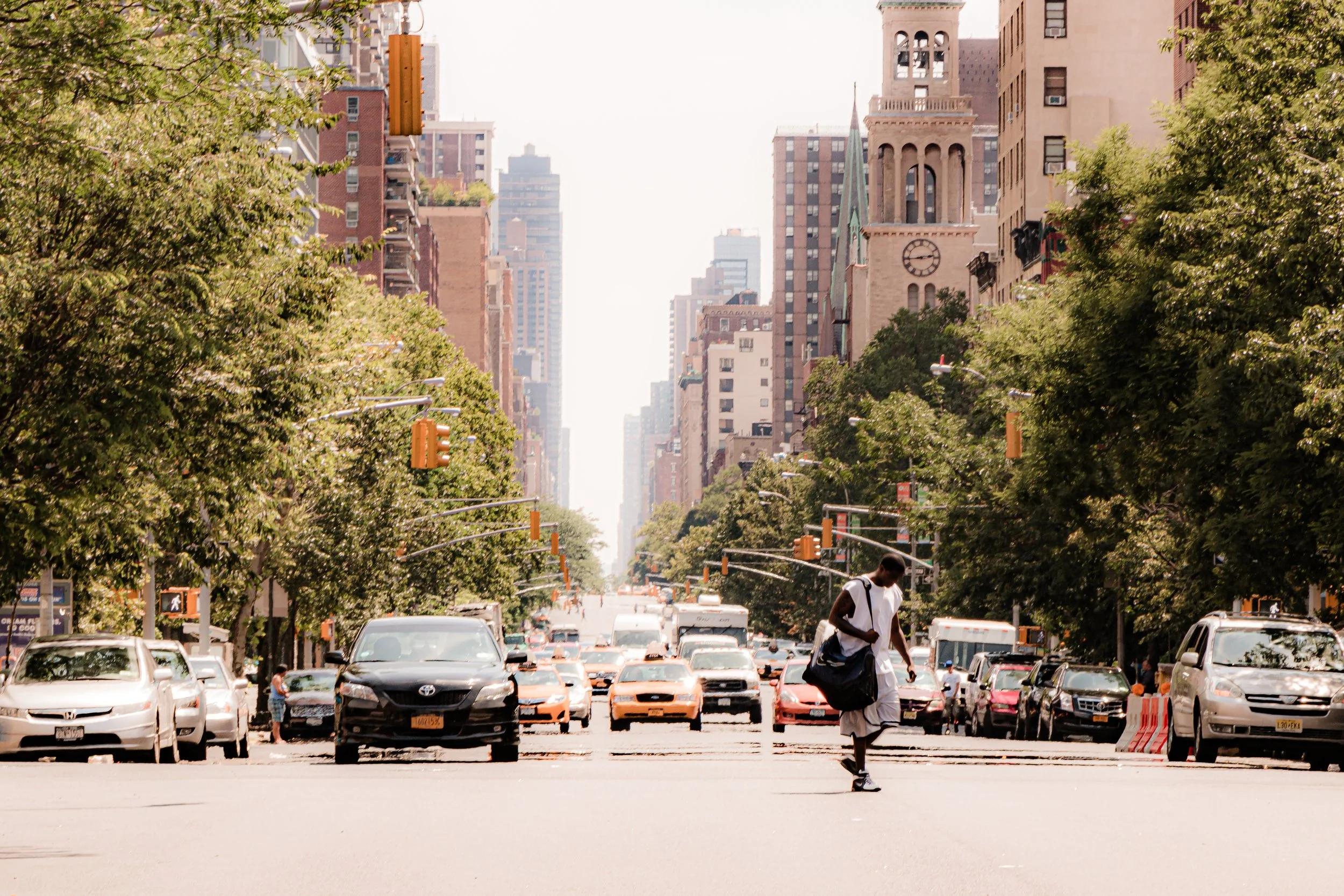 A city street scene with a man walking across the road carrying a bag, surrounded by cars and tall buildings, with green trees lining the street and a clock tower in the background.