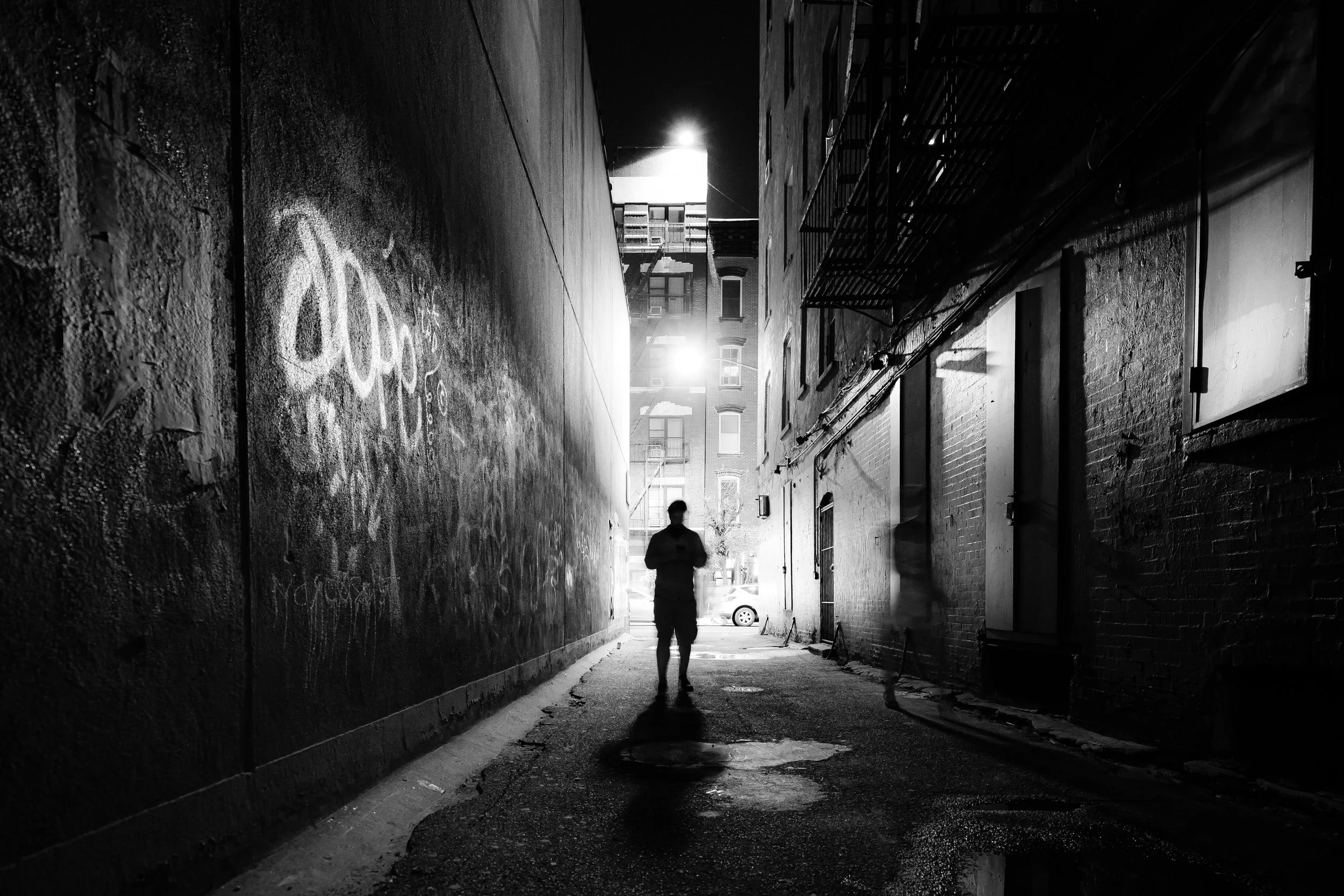 A black and white photo of a person walking through a narrow alleyway at night, with buildings on both sides, graffiti on the wall, and bright light illuminating the background.