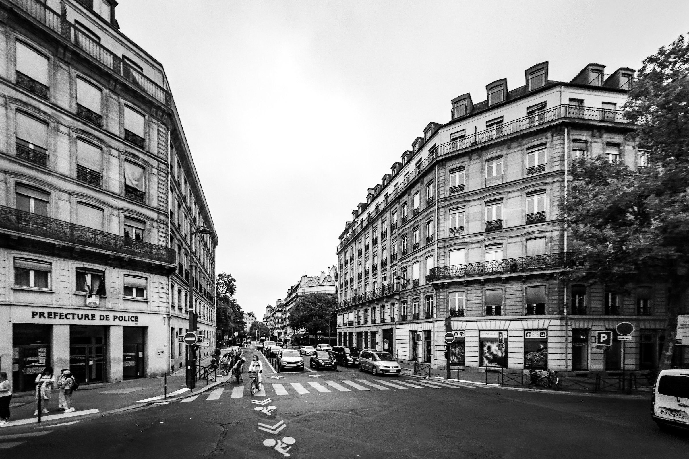 Black and white photograph of a Parisian street corner with classic Haussmann-style buildings, parked cars, bicycles, and pedestrians crossing a crosswalk. The building on the left bears a sign that reads 'Prefecture de Police'.