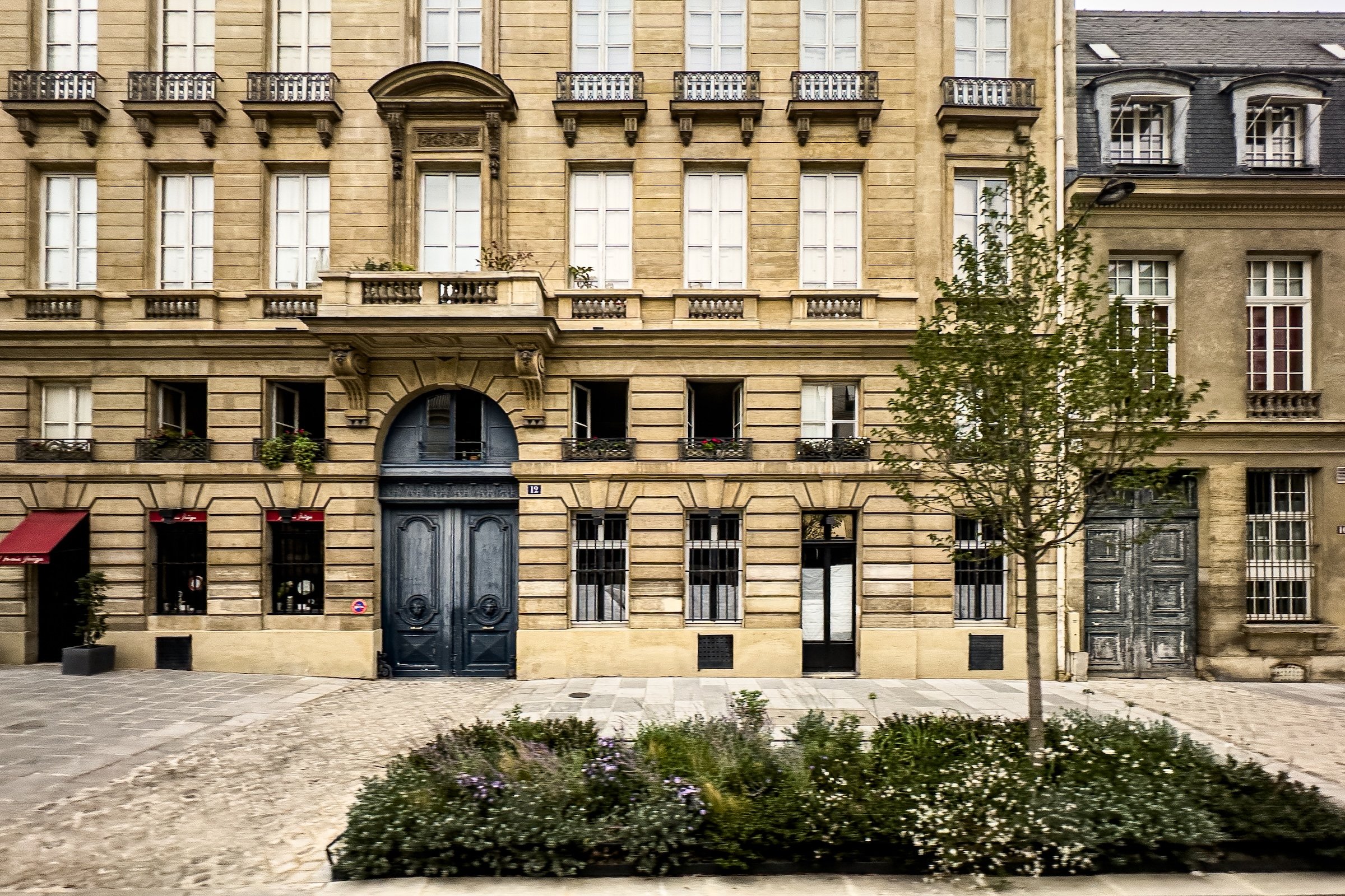 A street view of a classical European building with beige stone facade, tall windows, small balconies with iron railings, and a large arched entrance door.