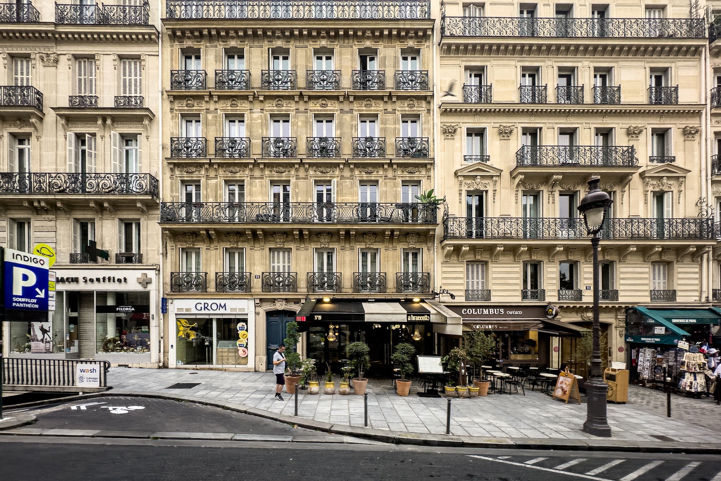 A busy Paris street scene with multi-story beige stone buildings featuring ornate wrought-iron balconies. The ground level has storefronts including a pharmacy, a cafe, and various shops. People are walking on the sidewalk, and a lamppost and street signs are visible in the foreground.