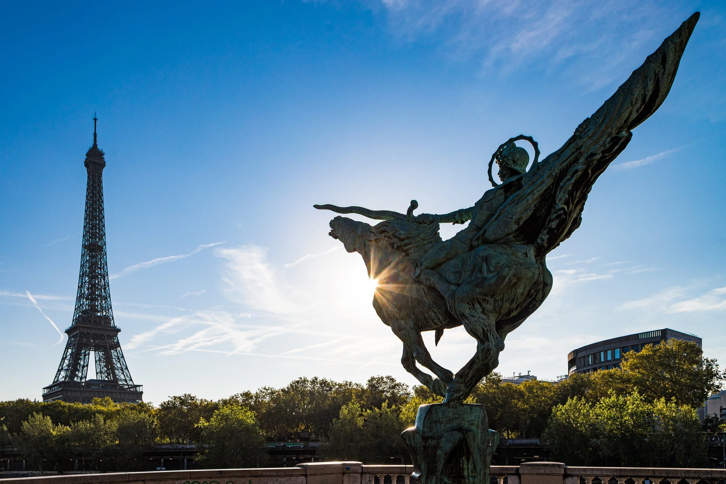 Statue of a winged figure with a sword, silhouetted against the sun with the Eiffel Tower in the background, in Paris.