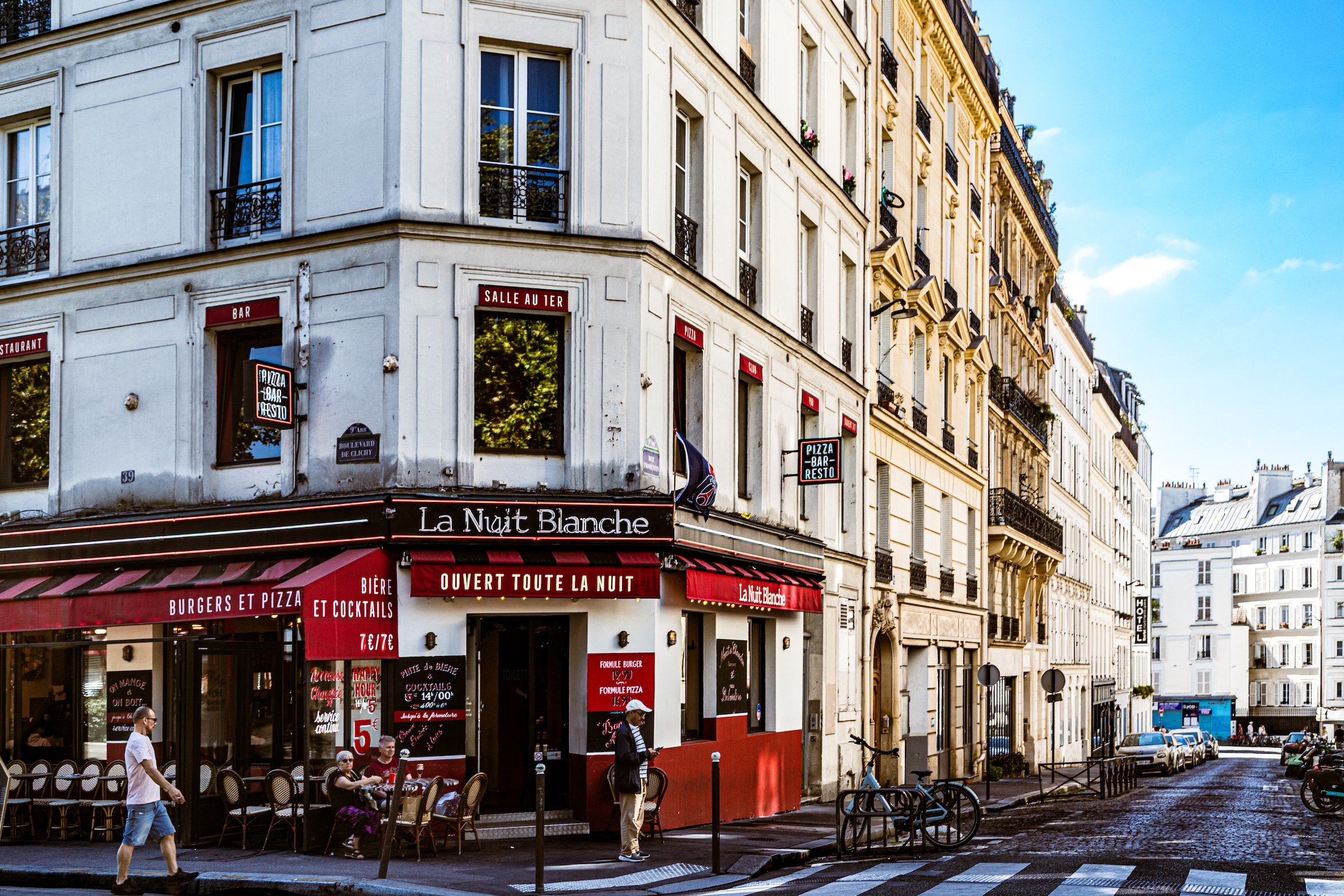 A street scene in Paris with a corner restaurant called La Nuit Blanche, featuring red and black signage and outdoor seating. The building has cream-colored walls, black wrought-iron balconies, and large windows. There are several pedestrians walking along the sidewalk, and bicycles are parked nearby. The sky is clear and blue.