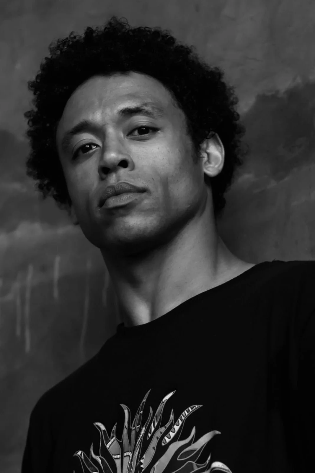 Black and white portrait of a person with curly hair, looking confidently at the camera, wearing a dark shirt with a graphic design.