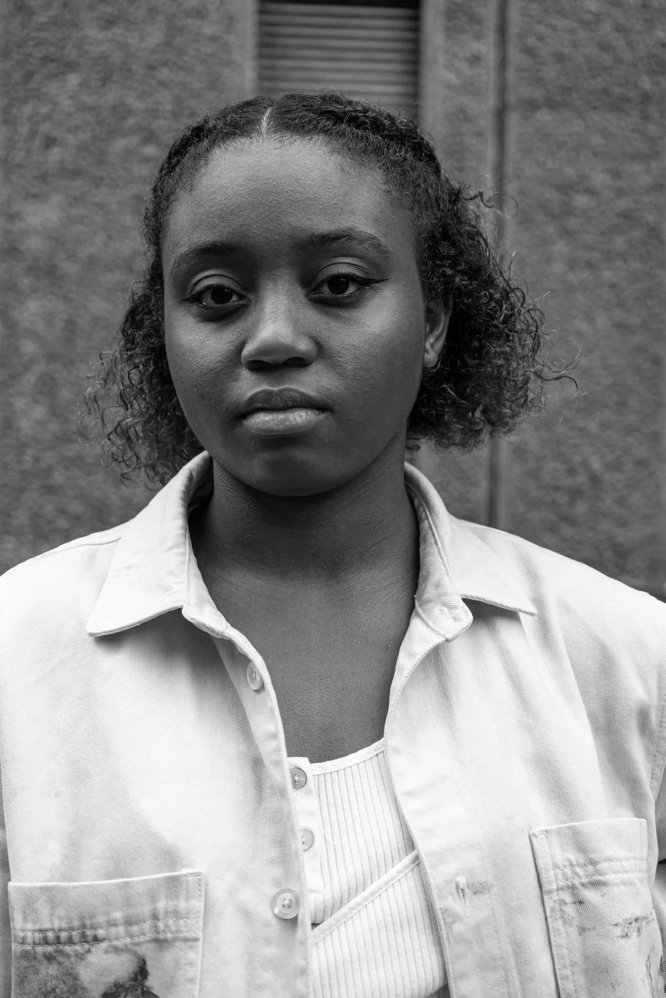 Black and white portrait of a woman with short curly hair, wearing a light-colored denim jacket over a ribbed shirt, standing outdoors in front of a textured wall.