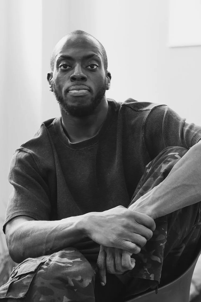 Black and white photo of a young man sitting, looking into the camera, wearing a dark t-shirt and camouflage pants.