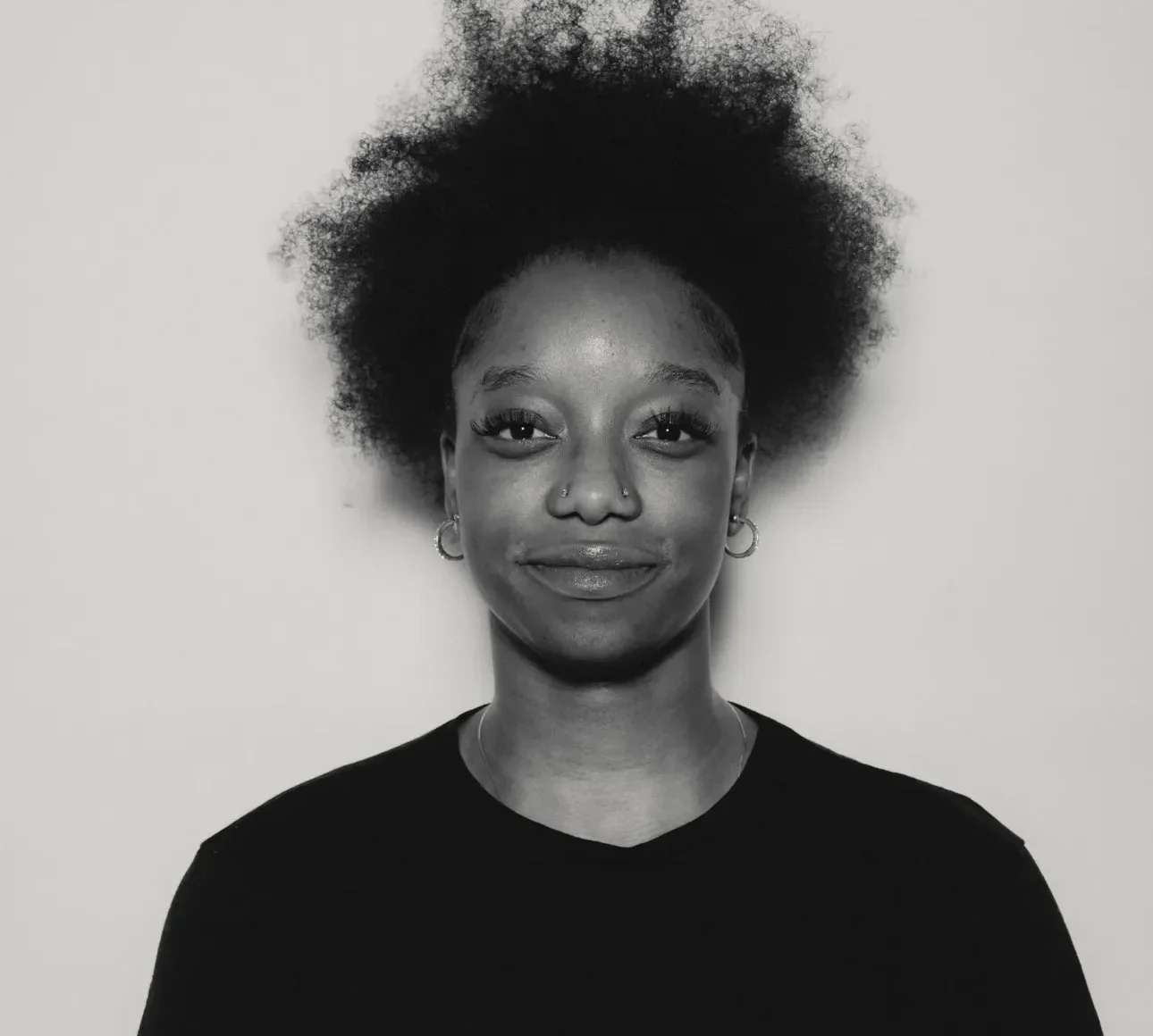 Black and white portrait of a young woman with natural afro hair, earrings, and a nose piercing, smiling softly against a plain background.