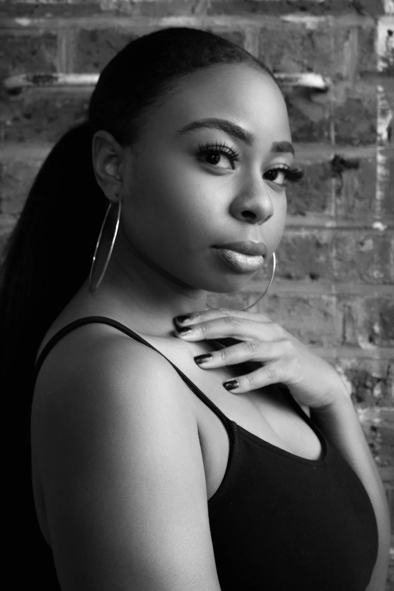 Black and white portrait of a young woman with hoop earrings, wearing a sleeveless top, looking at the camera with a confident expression, set against a brick wall background.