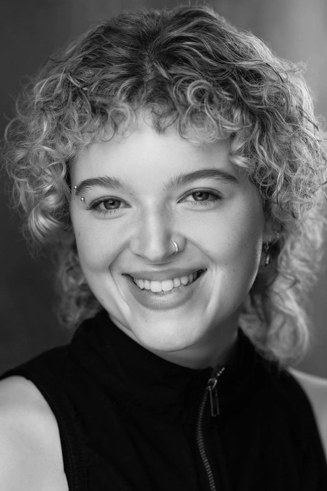 A smiling young woman with curly hair, piercing, and a nose ring, wearing a sleeveless black top, with a blurred background in black and white.