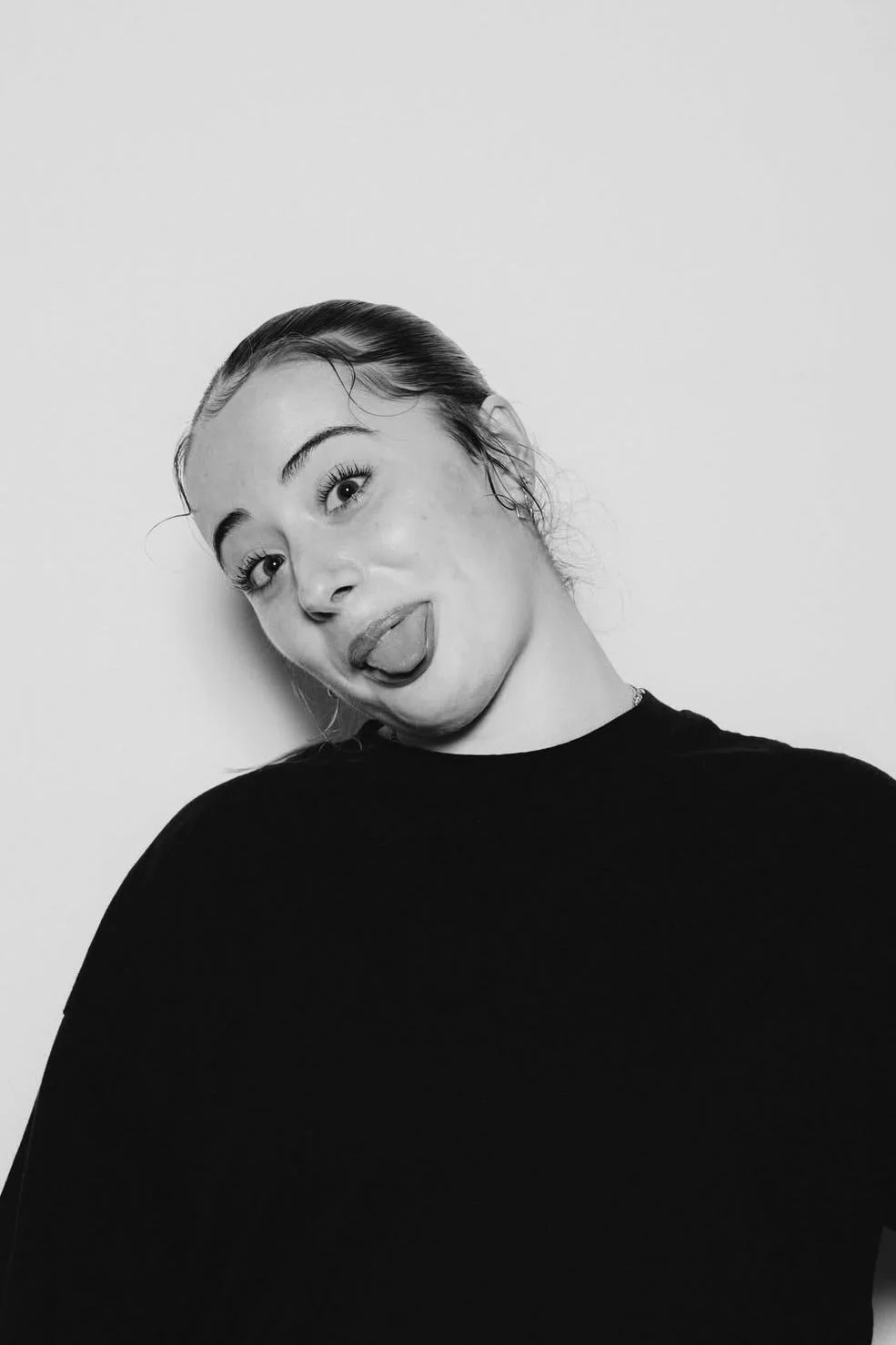 Black and white photo of a young woman sticking her tongue out and smiling slightly, with her head tilted to the side, wearing a dark shirt.