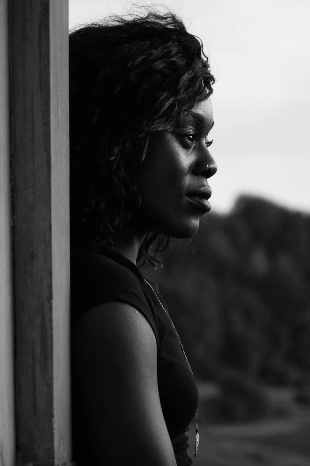 A black and white photograph of a woman with curly hair, standing outdoors, her side profile visible, looking contemplative.