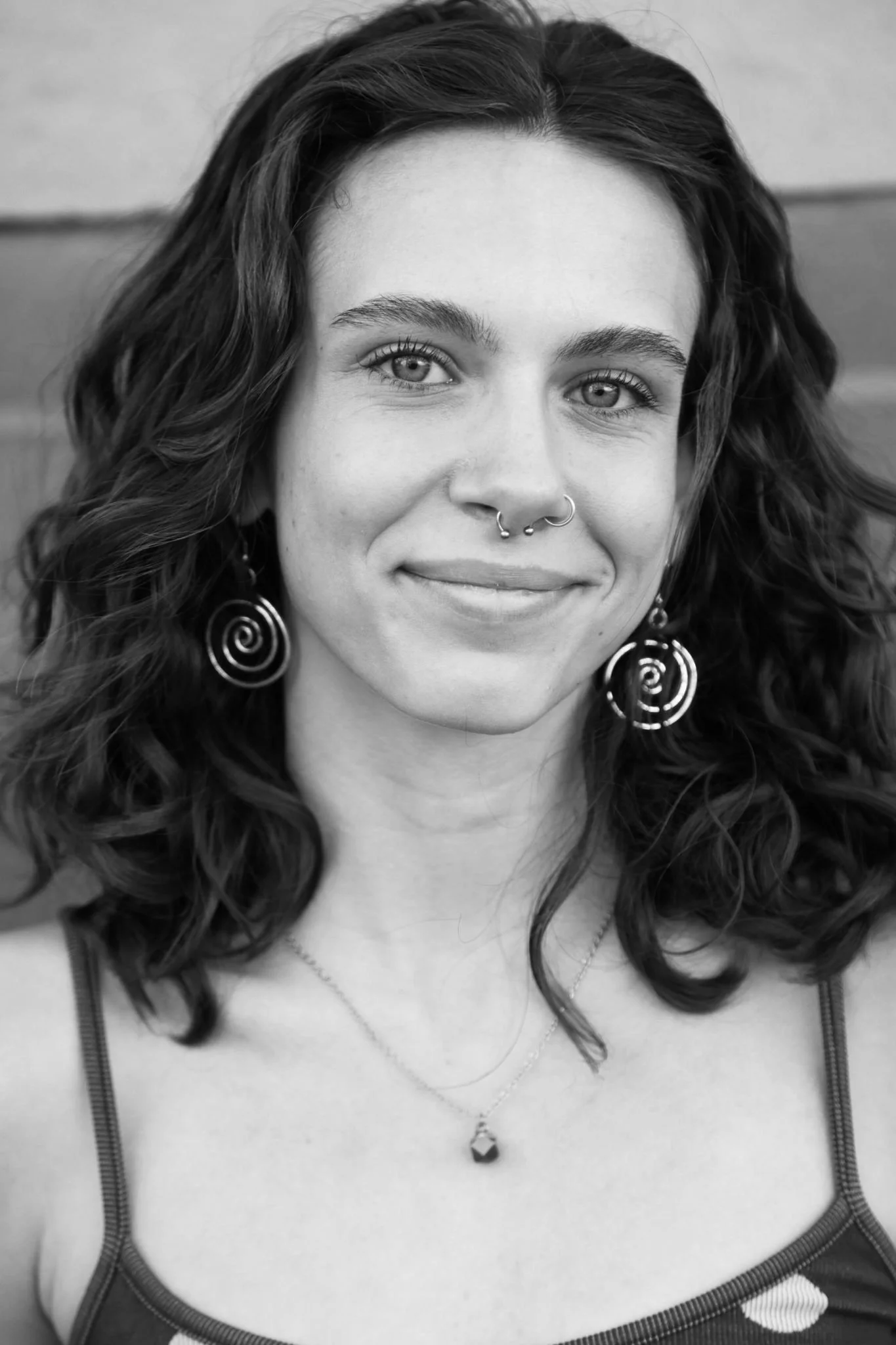 Black and white portrait of a woman with curly hair, earrings, and multiple facial piercings, smiling at the camera.