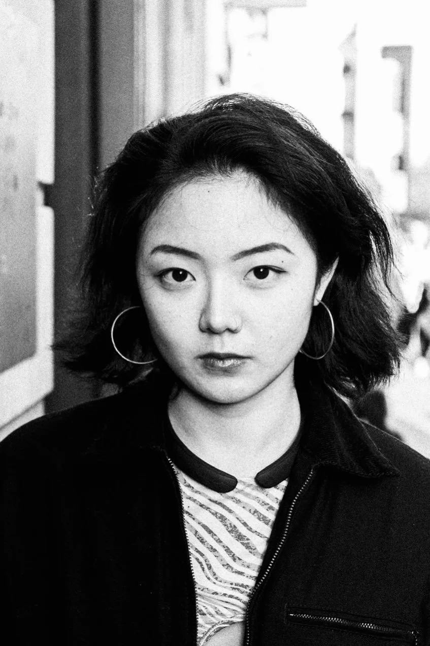 A black-and-white portrait of a woman with medium-length wavy hair, wearing hoop earrings and a zip-up jacket, standing outdoors.