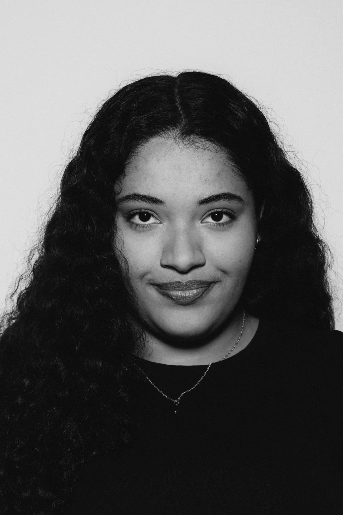 Black and white portrait of a woman with long, curly hair, smiling, wearing earrings, a necklace, and a dark top against a plain background.