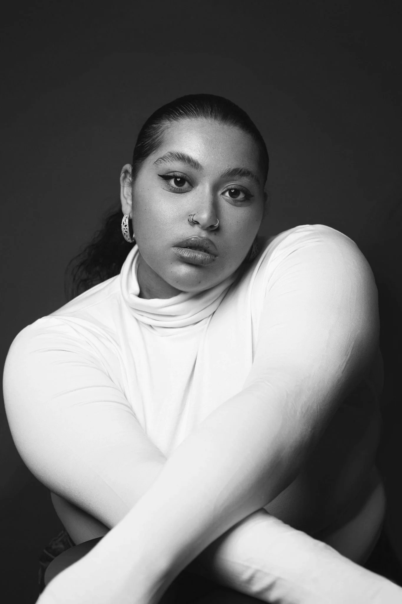 Black and white portrait of a young woman with dark hair pulled back, wearing hoop earrings, a nose ring, and a white high-neck top, posing against a dark background.