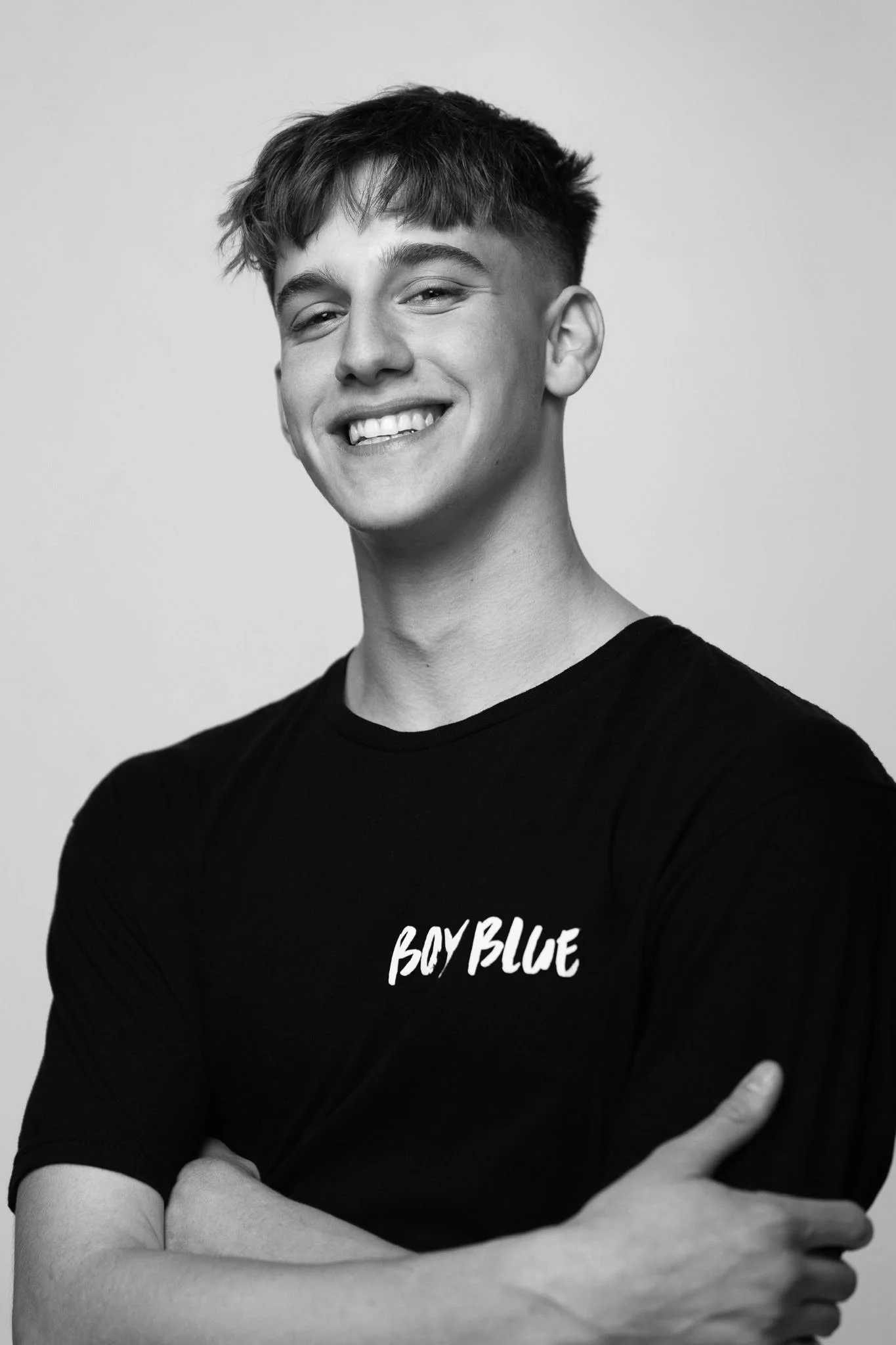 Black and white portrait of a young man with a big smile, wearing a black T-shirt that says "Boy Blue." He has short, tousled hair and his arms are crossed.