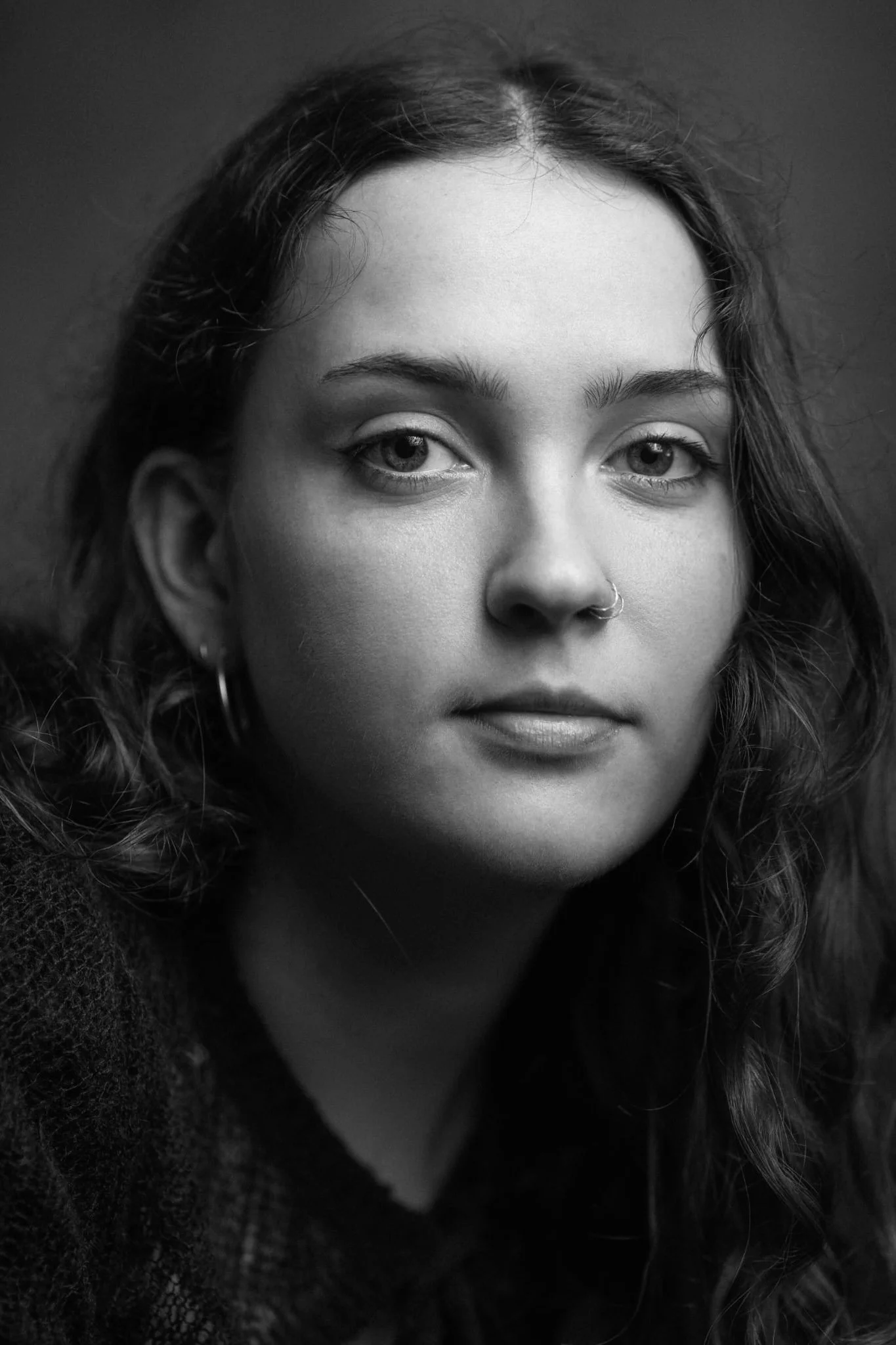 Close-up black and white portrait of a young woman with wavy hair and a nose ring, looking at the camera.